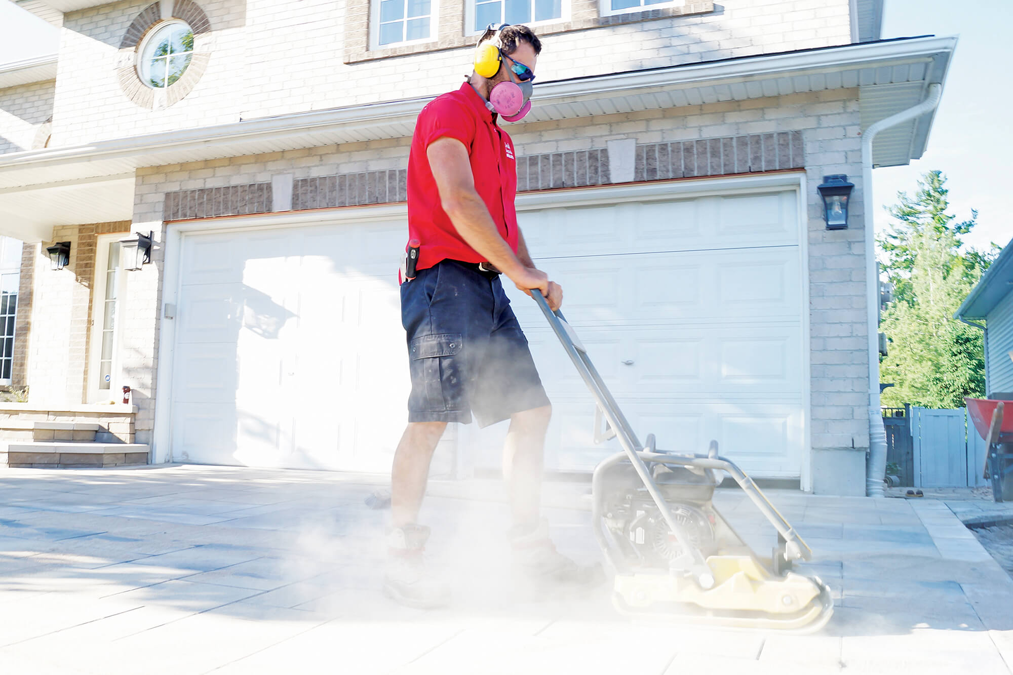 man wearing a mask using a plate compactor on a driveway with lots of dust