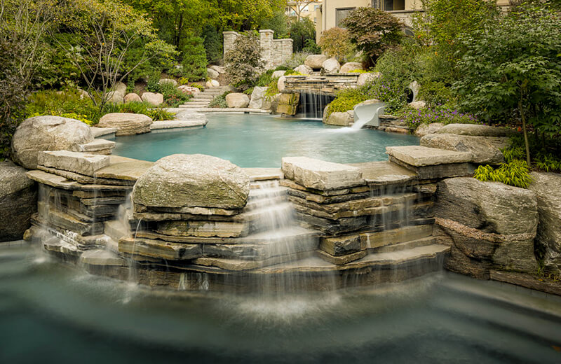 natural rock waterfall in a pool with a house in the distance