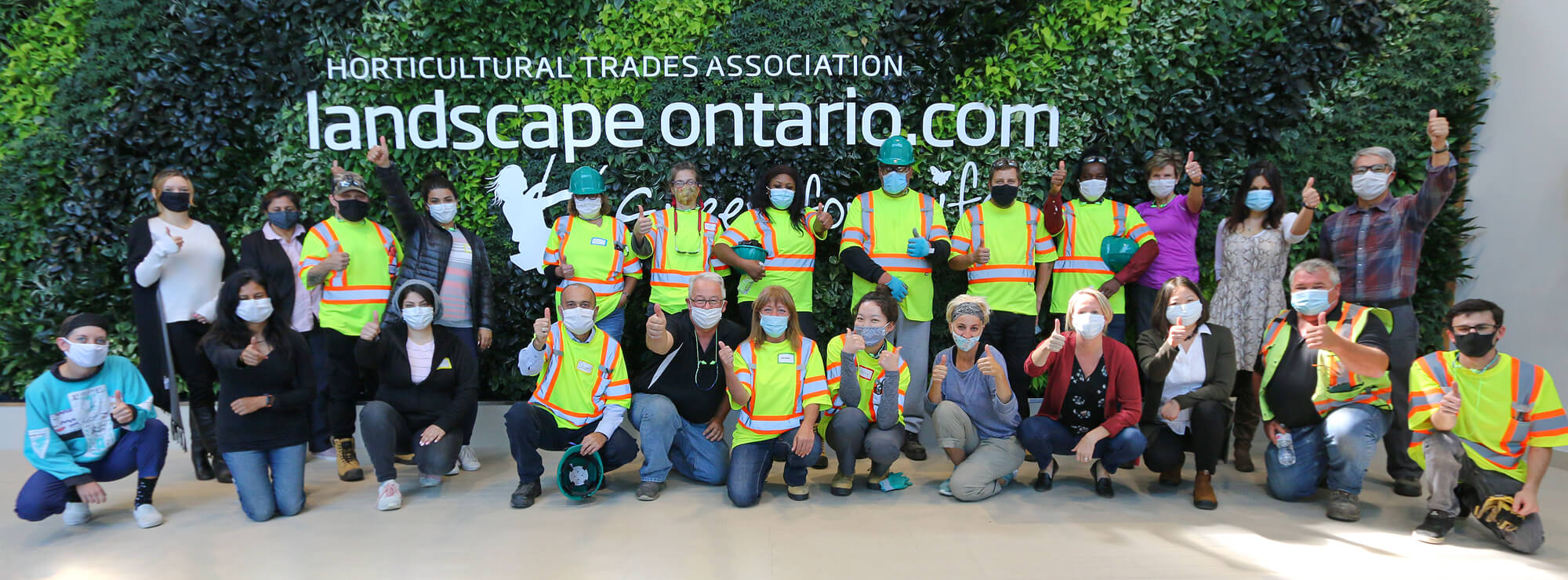 large group of people wearing PPE and face masks in front of a living wall