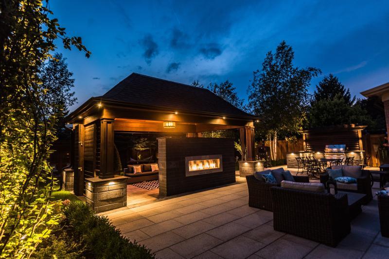 2018 - Landscape Lighting Design & Installation - Under $10,000 - This photo captures all of the lighting in the backyard. Pot lights install along the overhang of the pavilion, strip light install underneath all copping of the pillars and counter top of the outdoor kitchen, and uplight fixtures inside the different planting bed around the backyard