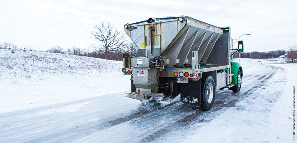 A large snow plow with a salt spreader on the back driving down a windy road in winter