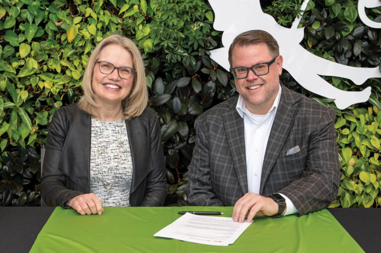 a blond woman wearing glass with a man in a suit jacket and glasses, both sitting at a table with a plant wall in the background